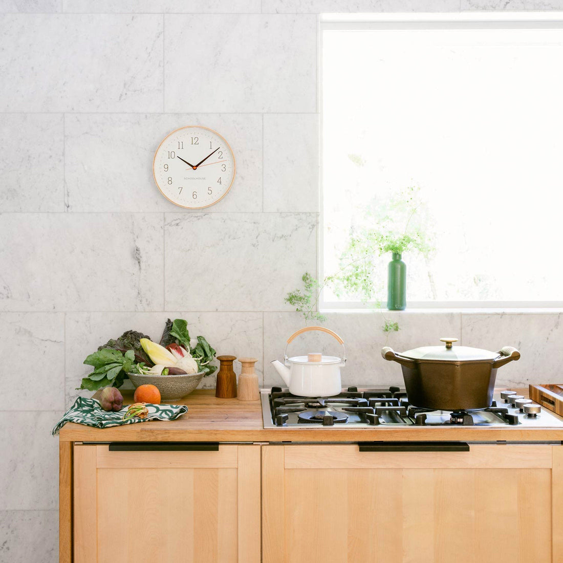 Round wall clock above a light-wood kitchen counter with stovetop, pot, kettle, and a bowl of produce by a bright window