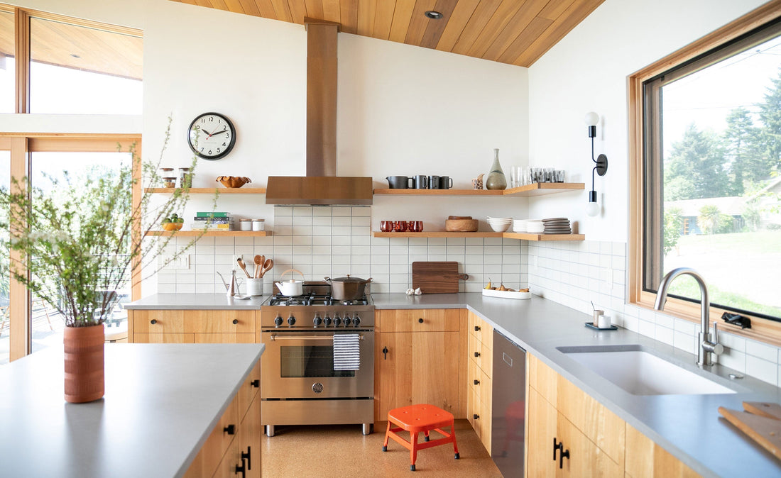 Wall clock mounted above open shelves in a bright modern kitchen with wood cabinets, stainless range, and large window
