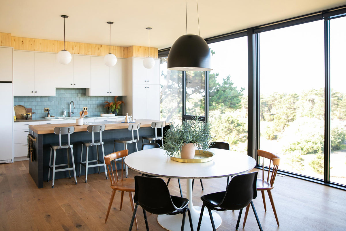 Open kitchen and dining area with round table and plant, wood breakfast bar with metal stools, pendant lights, large windows