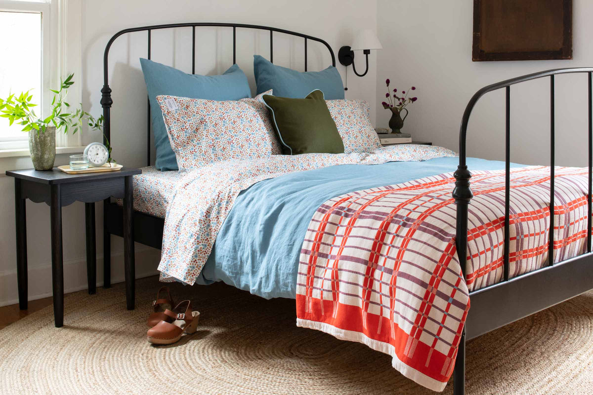 Bed dressed in floral sheets and a blue linen duvet with an orange plaid throw in a sunlit bedroom with a jute rug and nightstand