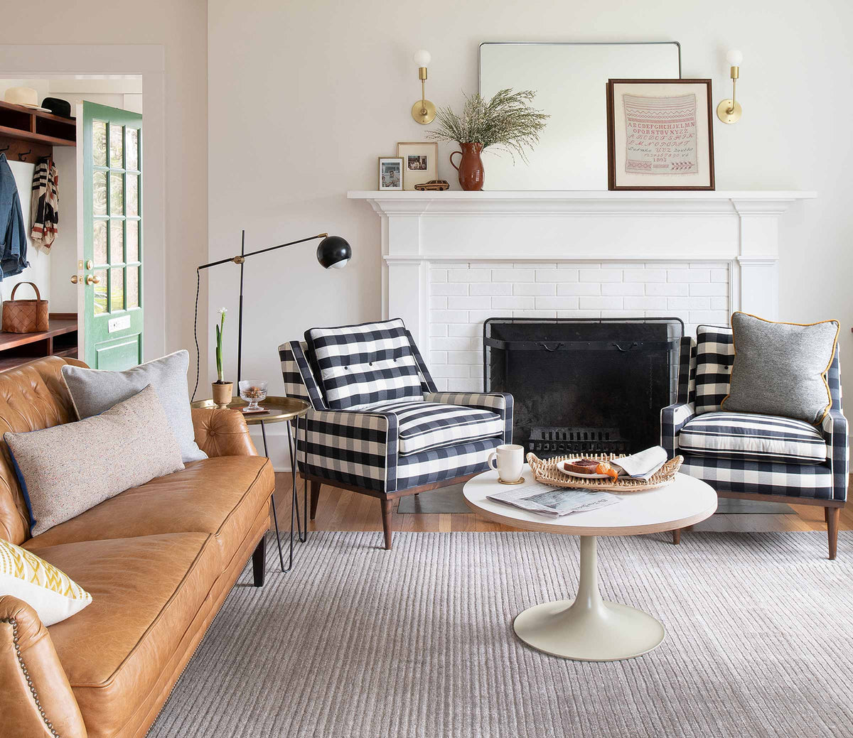 Living room with tan leather sofa and two black-and-white buffalo check armchairs facing a white fireplace, round pedestal coffee table and rug
