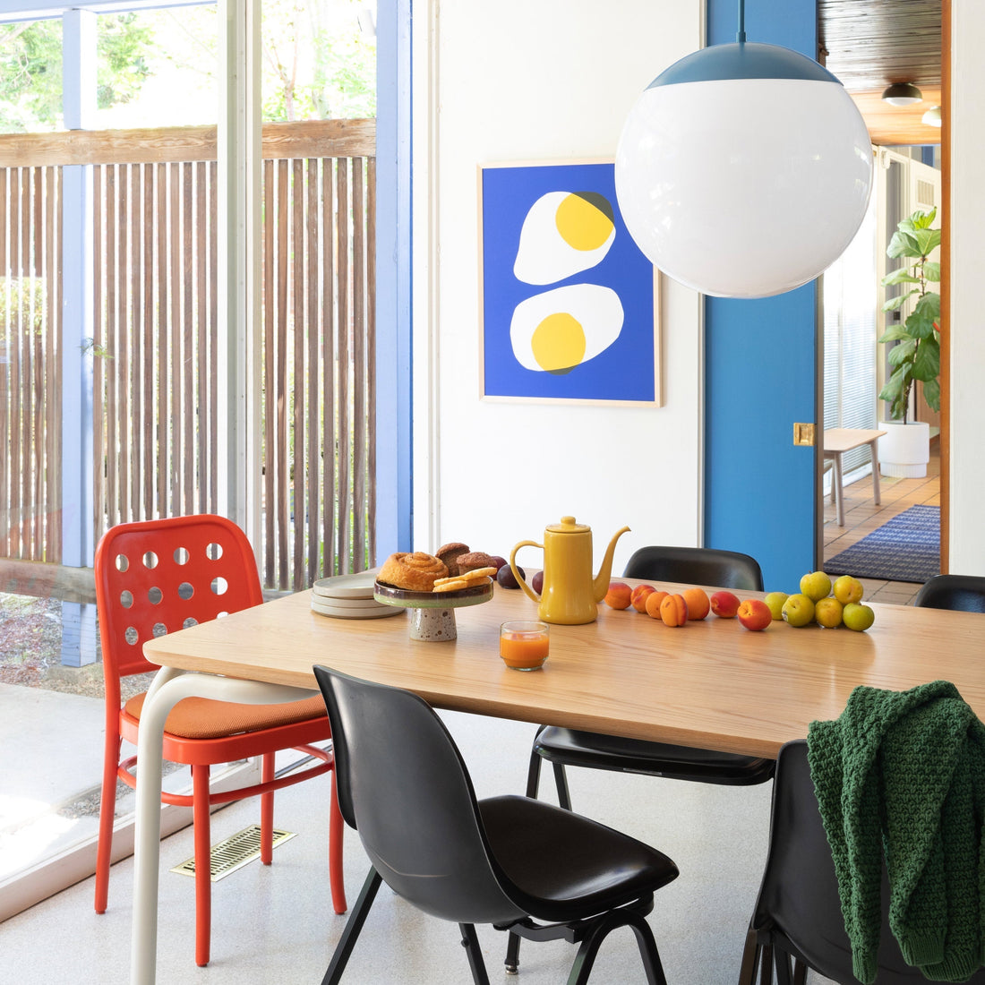 Dining table set for casual breakfast with globe pendant, yellow teapot, pastries and fruit in a bright midcentury room