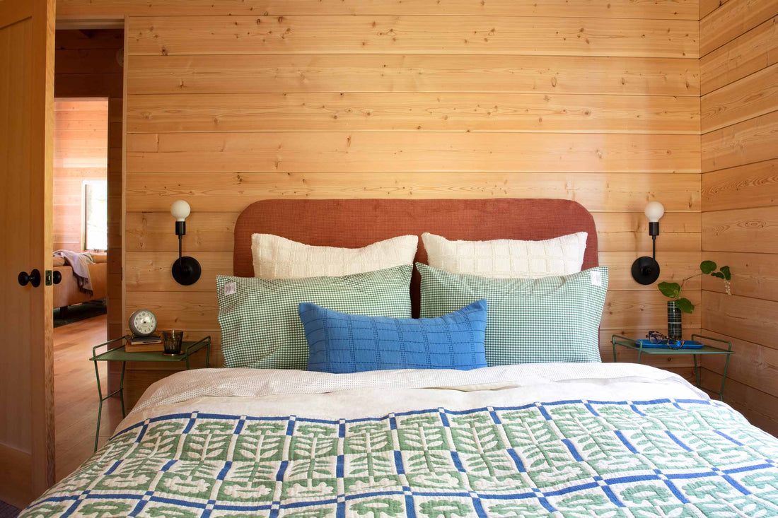 Bed with green-and-blue quilt, green gingham shams and blue lumbar pillow against a rust headboard in a wood-paneled room