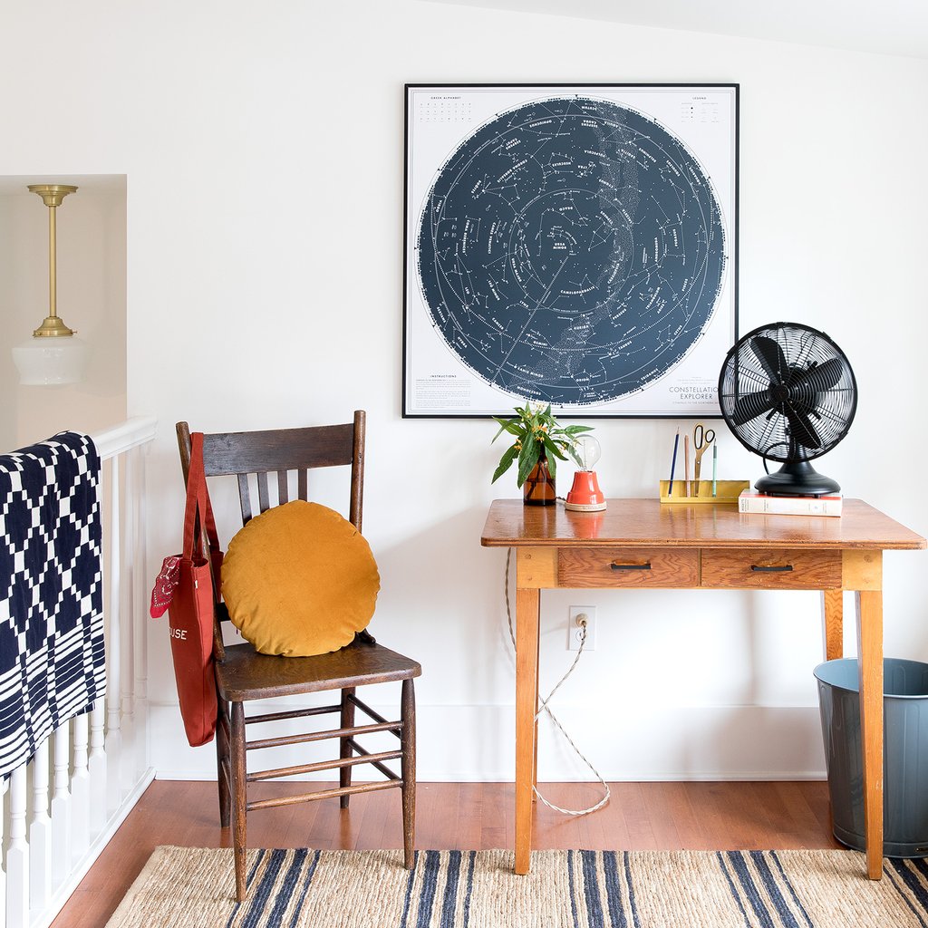 Wooden desk and chair styled with a framed star map, amber plant vase, black tabletop fan, and mustard round pillow in a bright landing nook