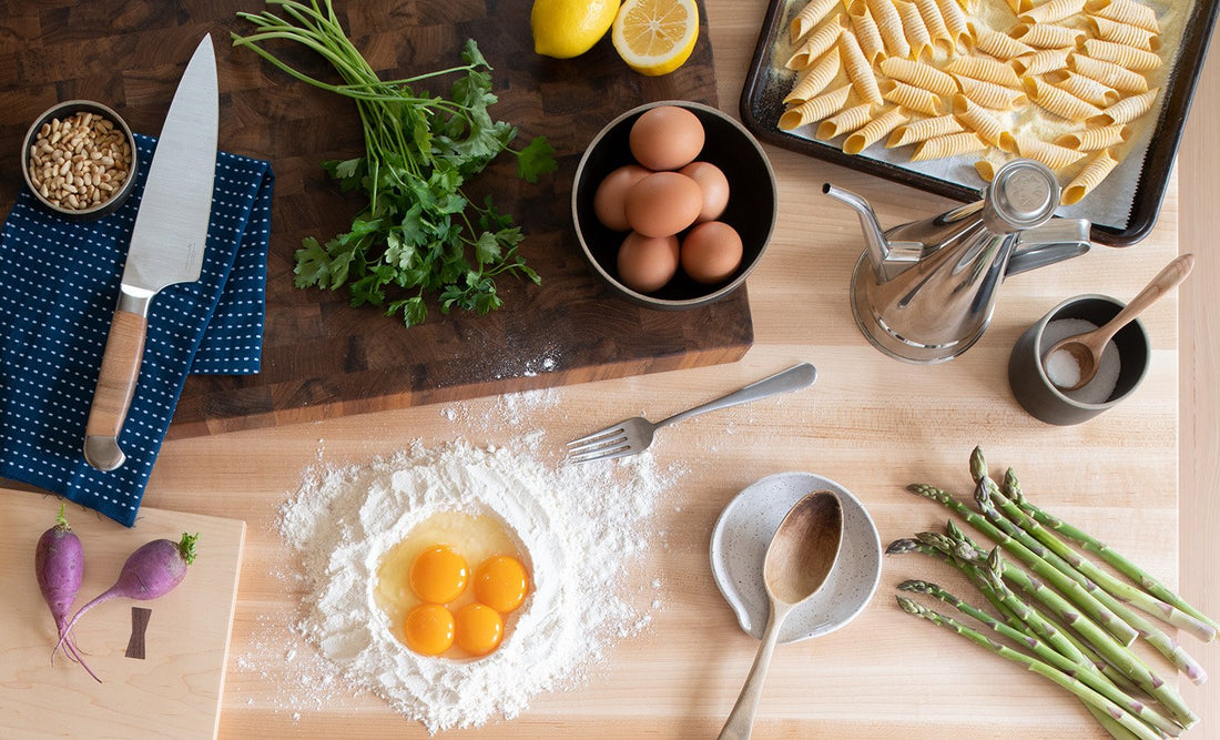 Ingredients for pasta primavera on a wooden counter: egg yolks in a flour well, eggs in a bowl, fresh asparagus, parsley, knife, wooden spoon, and sheet pasta