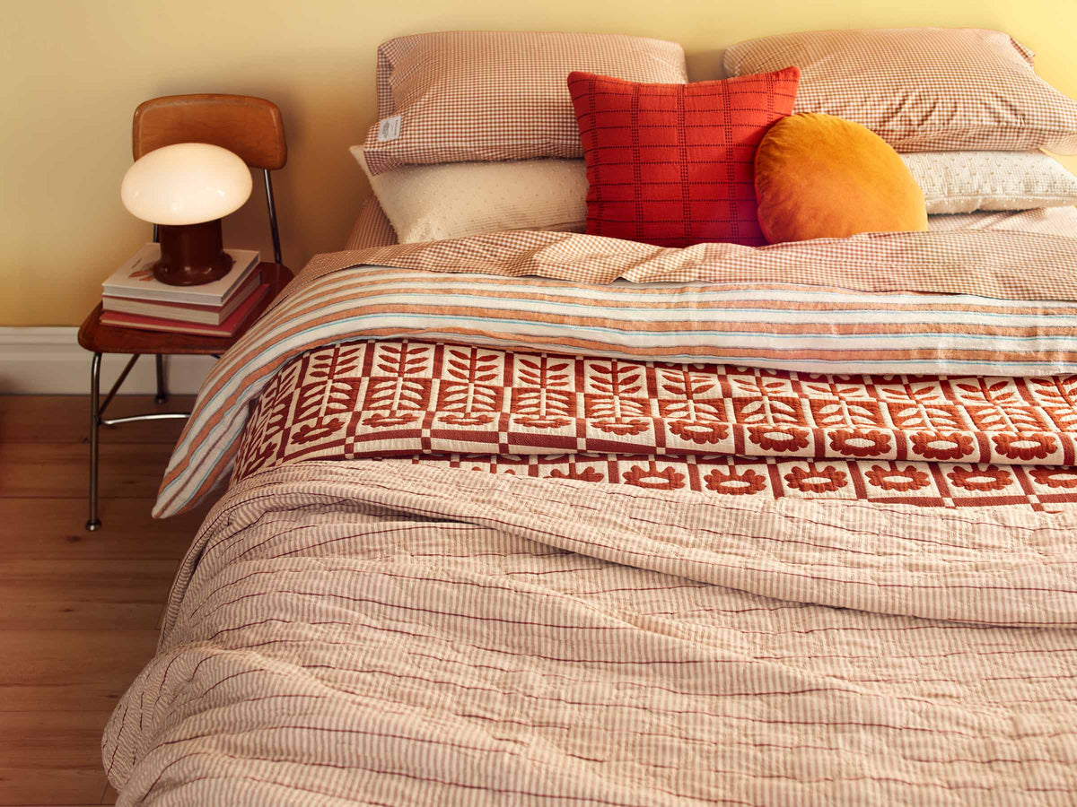 Bed layered with patterned quilts and gingham pillows, accented by rust-red and mustard cushions beside a wooden side table and lamp
