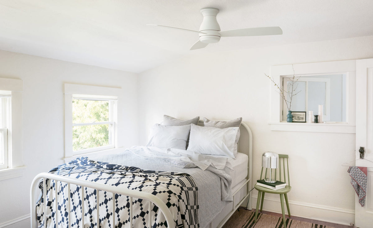 White ceiling fan above a sunlit small bedroom with a white metal bed, blue patterned quilt, and green side chair
