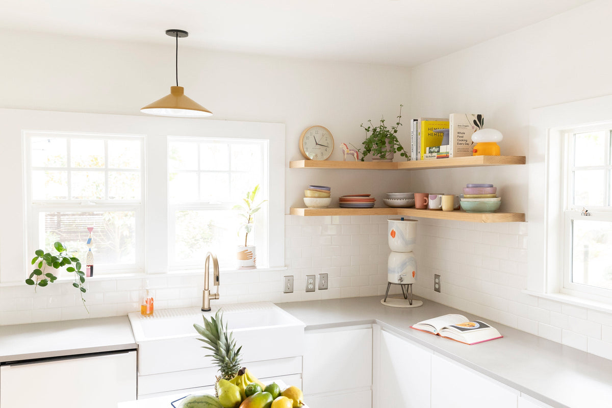 Sunlit white kitchen with brass pendant light over sink, open wood shelves holding colorful dishes and potted plants, fruit bowl on island