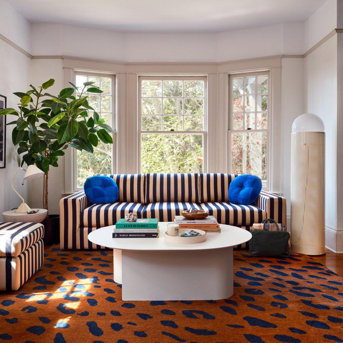 Striped navy and beige sofa with blue pillows in sunlit bay-window room with white coffee table and orange patterned rug
