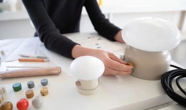 Person adjusting a finish knob on mushroom-shaped table lamps on a workshop bench with finish samples and sketches