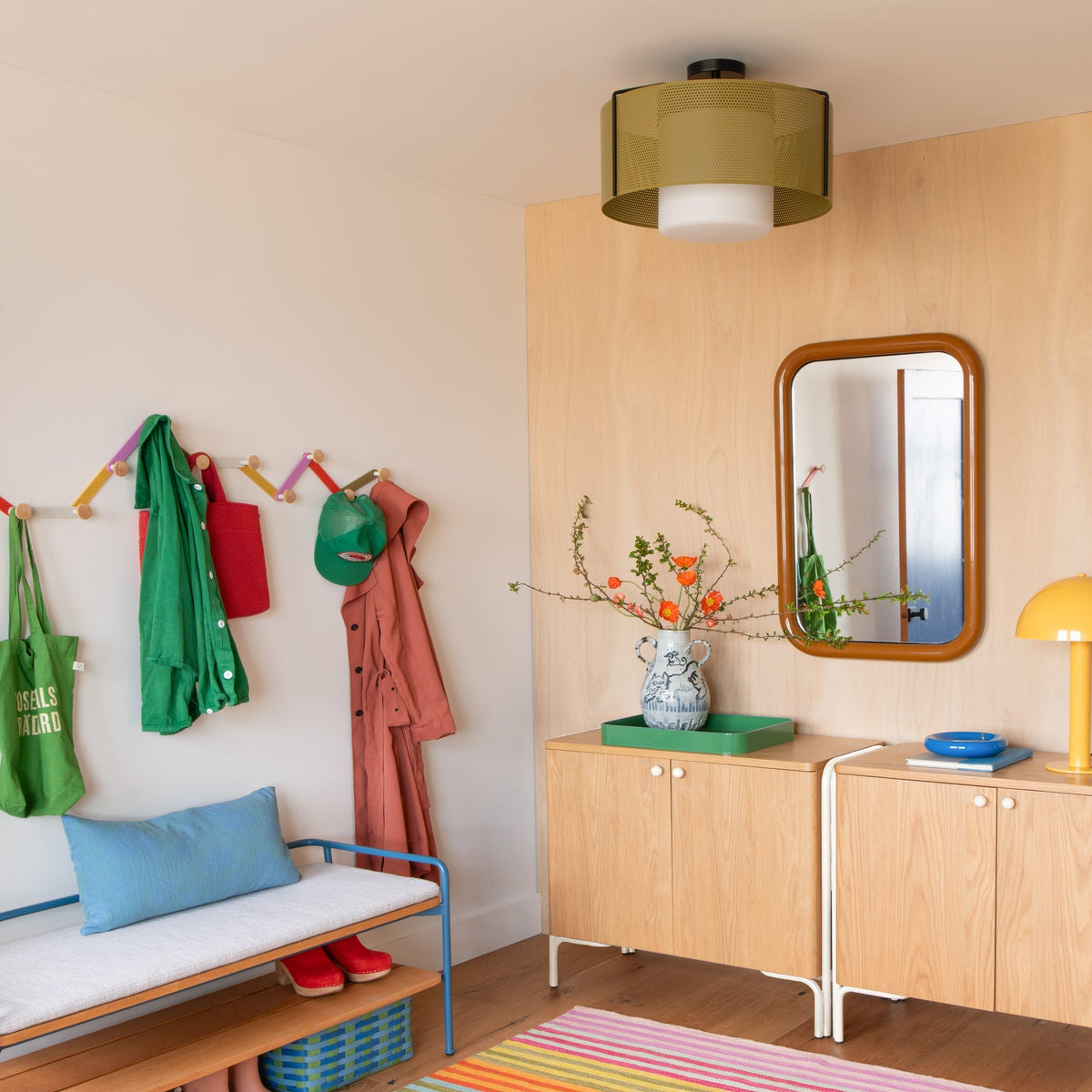 Tula hooks hold coats and hats on a colorful wall in a warm entryway with a bench and wood cabinets