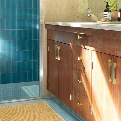 Angled view of Roland cabinet knobs in natural brass on a dark wood bathroom vanity with blue tile