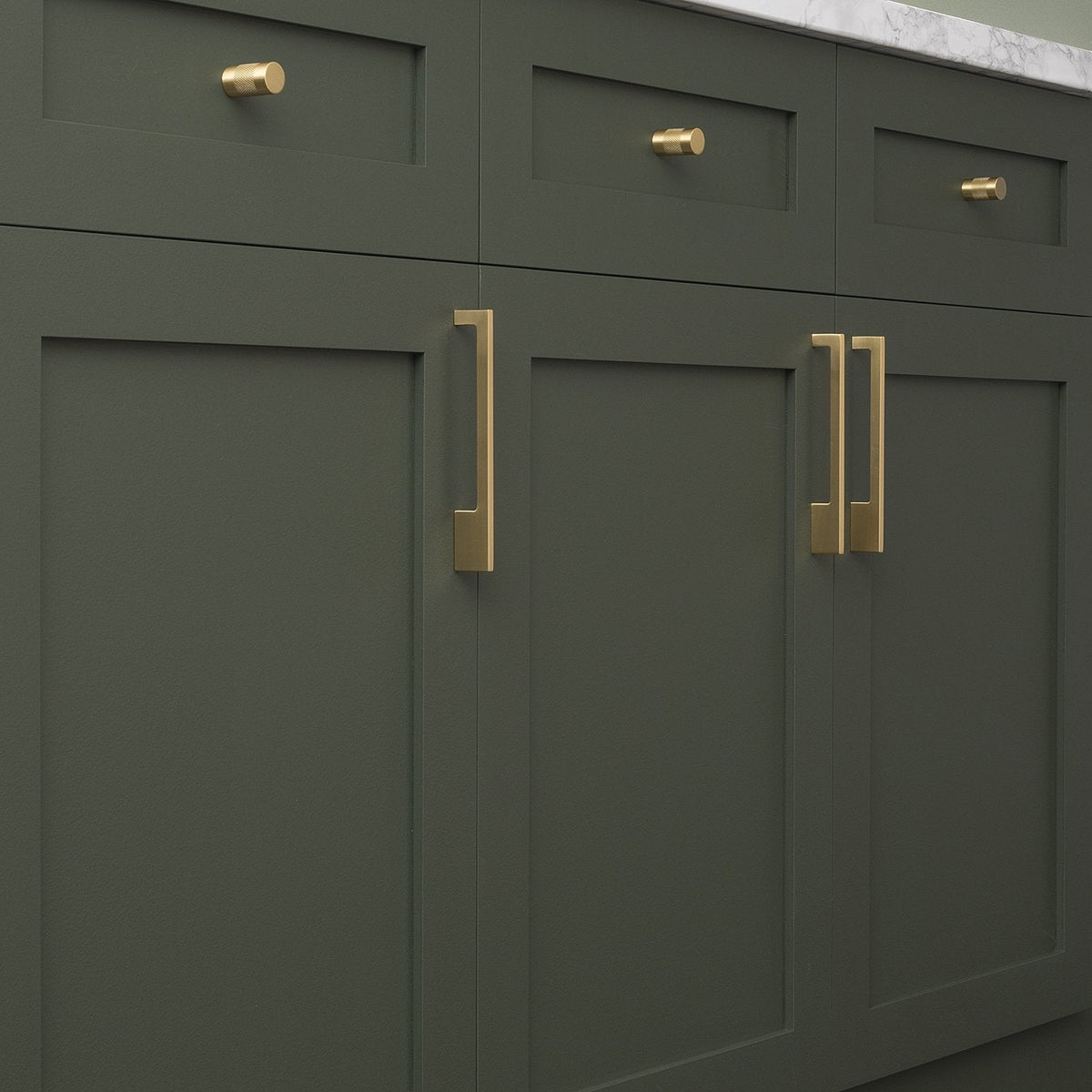 Natural brass knurled knobs adorn dark green kitchen cabinets beneath a marble countertop