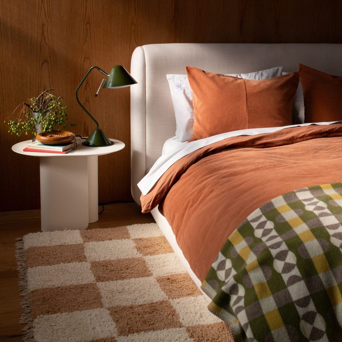 Yves cast metal catch-all on a white bedside table beside a green lamp in a bedroom vignette with rust-colored bedding and wood-paneled wall