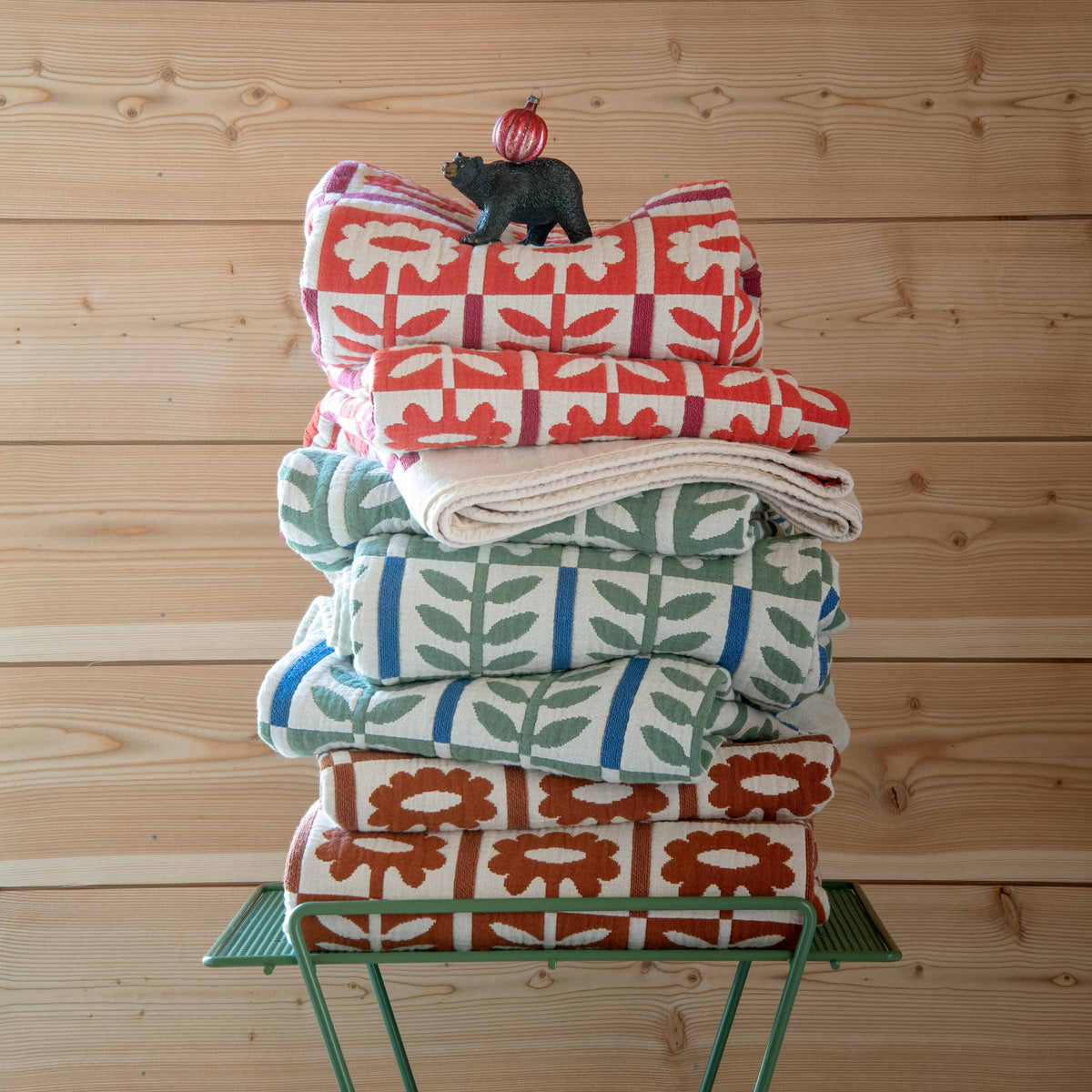 Front view of stacked Stillwater Floral Quilts showcasing multicolor botanical patterns on a green side table against wood paneling